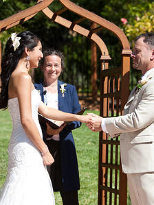 Corrina McFarlane officiating a garden wedding ceremony, couple holding hands under a wooden arbor