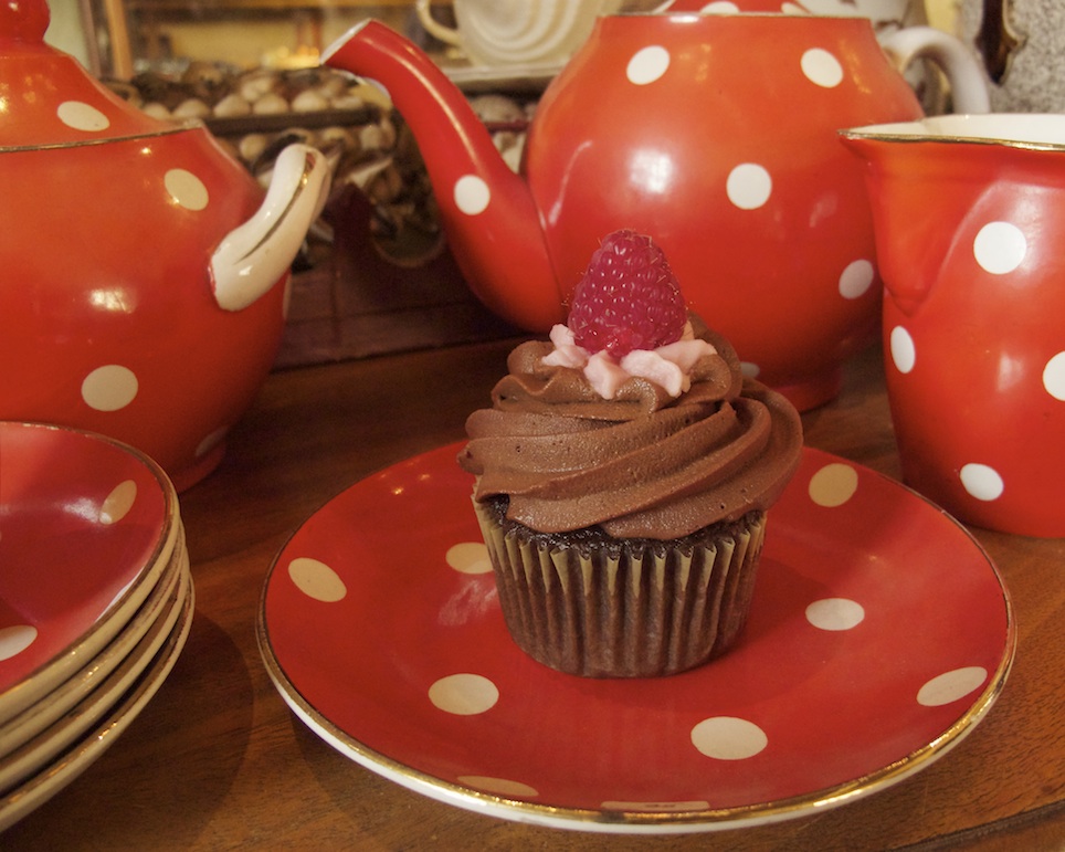 A chocolate raspberry cupcake on a red polka-dot plate with a matching tea set