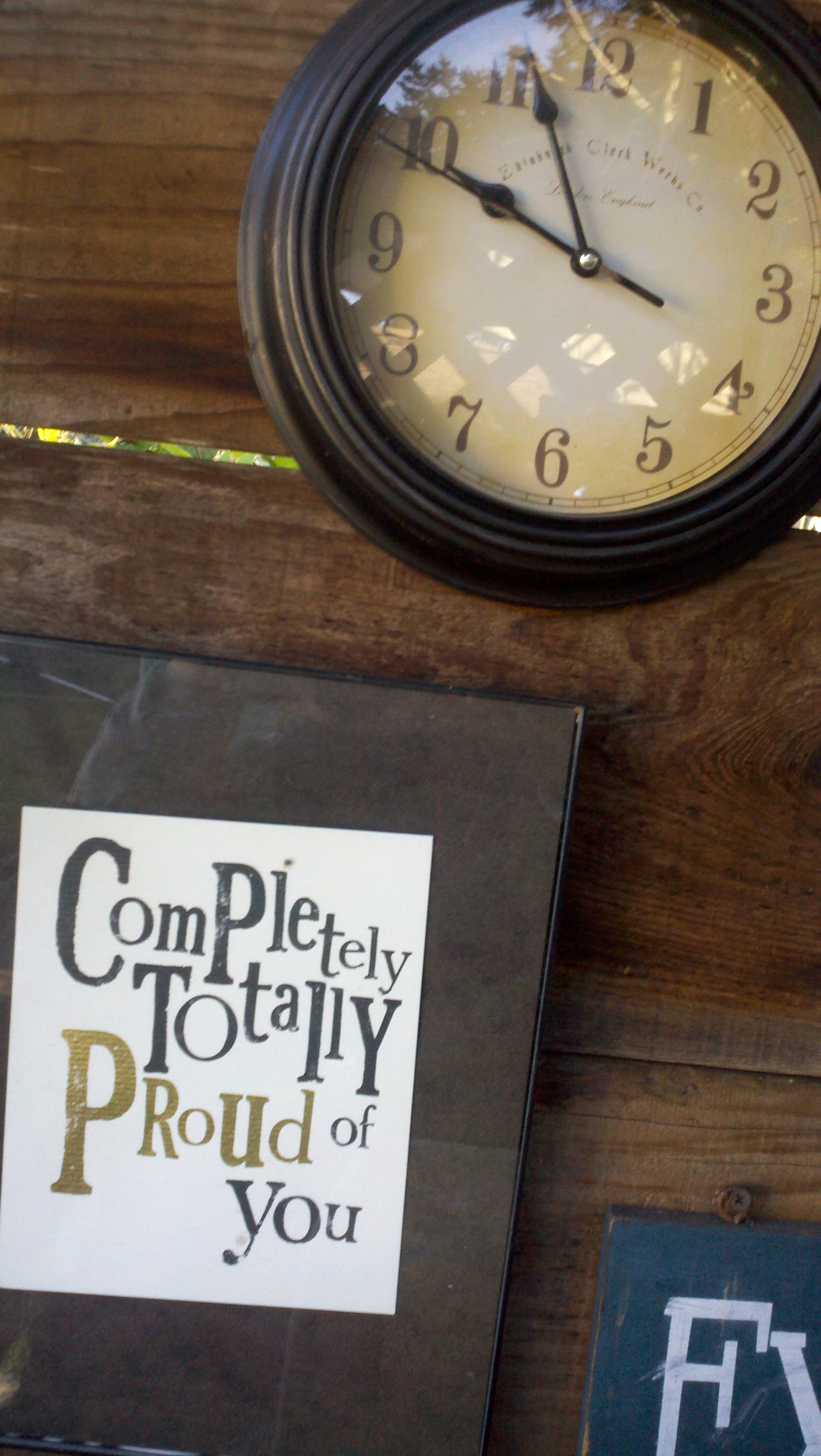 A framed print reading Completely Totally Proud of You beside a round clock on a wooden table